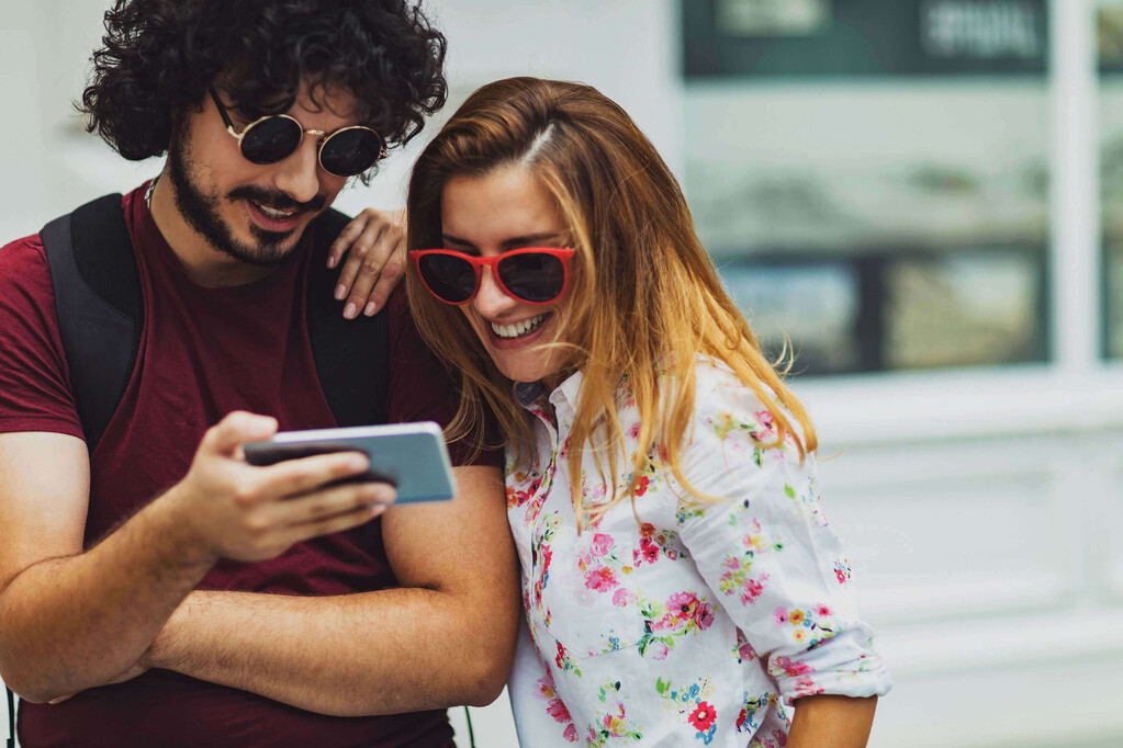 Junge Frau und Mann lächeln beim Blick aufs Smartphone – modernes Newsletterbildmotiv.